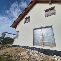 FIRMA USŁUGOWA - Świeżo otynkowana elewacja domu jednorodzinnego z drewnianą stolarką okienną, okna zabezpieczone folią, widoczne rusztowanie i pozostałości materiałów budowlanych na ziemi.