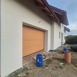 FIRMA USŁUGOWA - Świeżo otynkowana biała elewacja domu z brązowym dachem, widoczna drewniana brama garażowa, niebieskie beczki i elementy budowlane na niezagospodarowanym terenie.