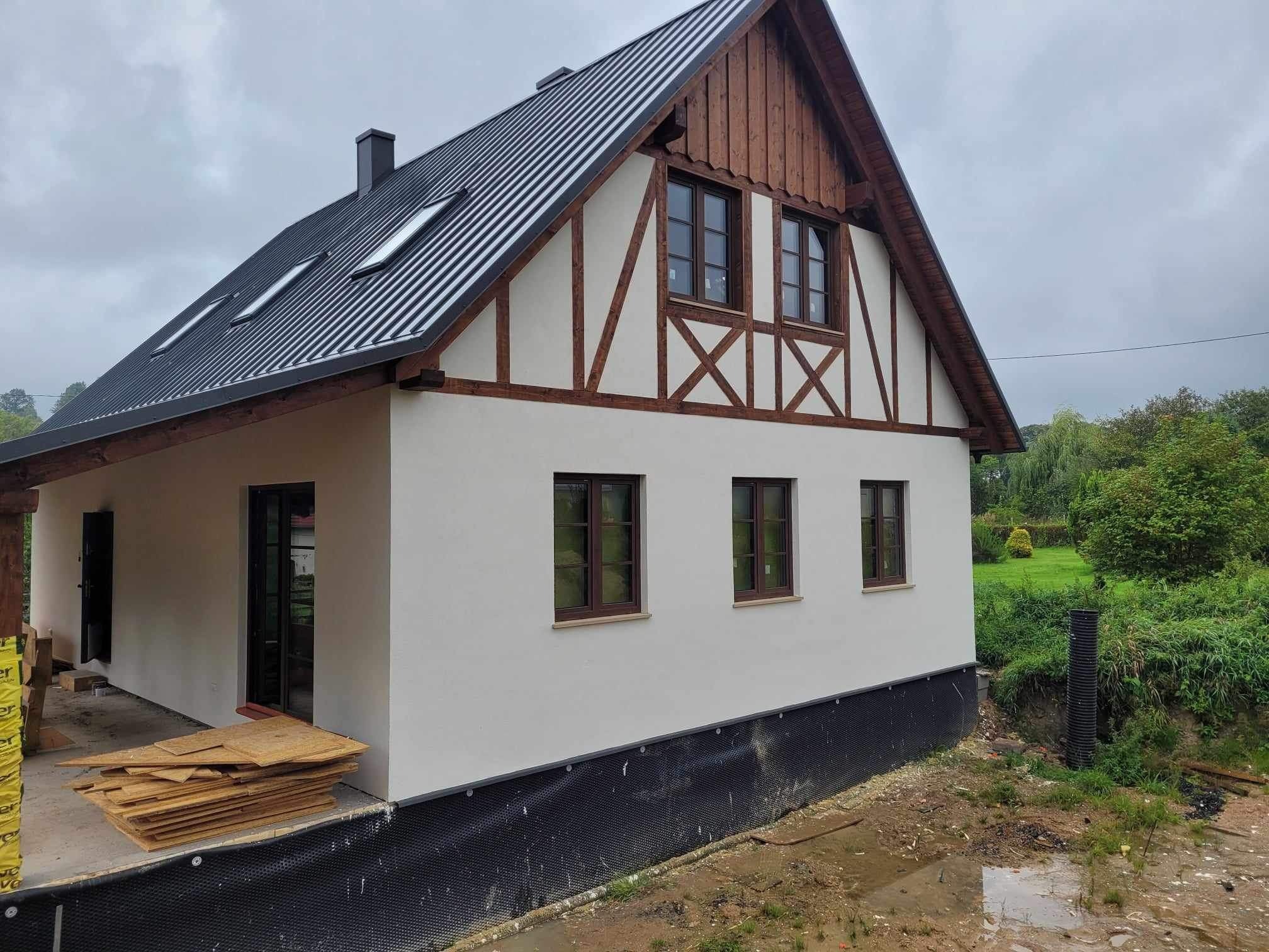 Nowy dom z jasną elewacją, drewnianą konstrukcją szkieletową i czarnym dachem z blachy trapezowej, widoczne okna dachowe i stos materiałów budowlanych przed wejściem.