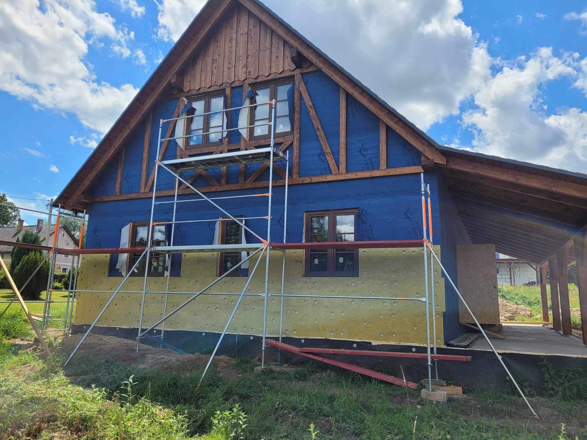 Dom w trakcie ocieplania, widoczne rusztowanie, niebieska membrana wiatroizolacyjna i żółta wełna mineralna, drewniana konstrukcja dachu, błękitne niebo z chmurami w tle.