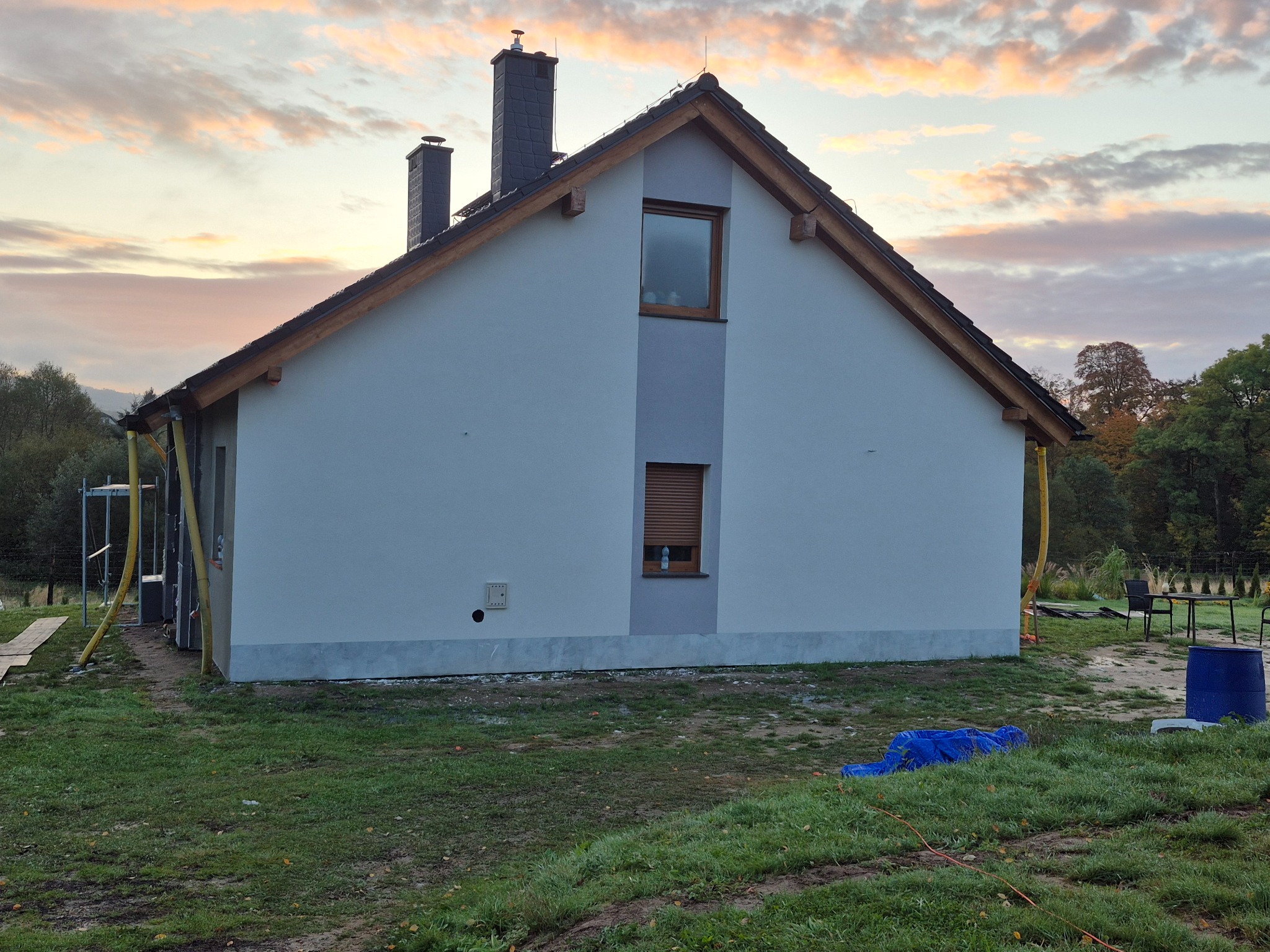 Świeżo otynkowany dom jednorodzinny z dachem pokrytym dachówką, widoczne rusztowania i elementy wykończeniowe, krajobraz jesienny w tle.