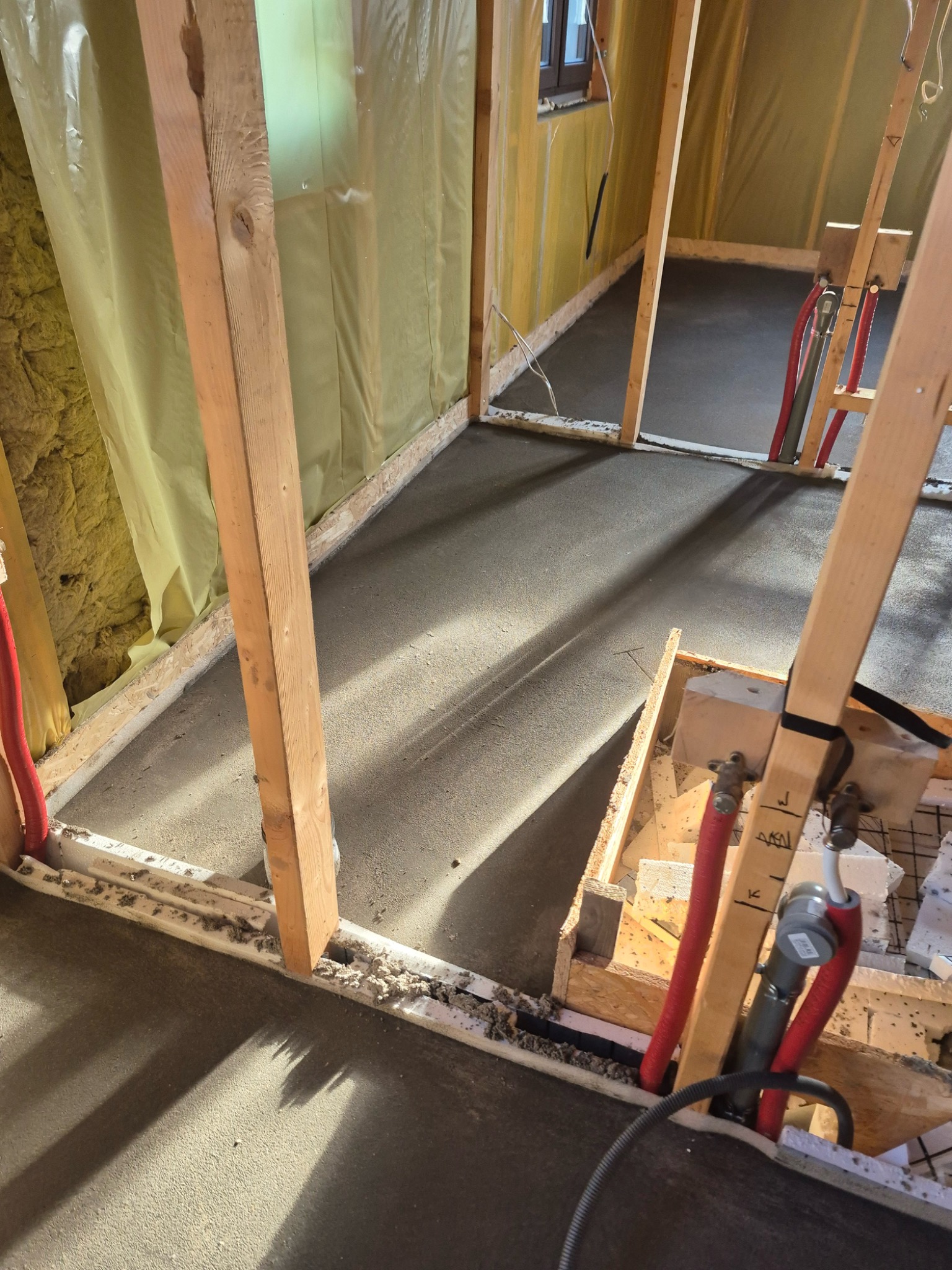 Wylewka betonowa na podłodze w trakcie budowy, widoczne drewniane ramy konstrukcyjne, izolacja ścian wełną mineralną i rury instalacji grzewczej podłogowej.