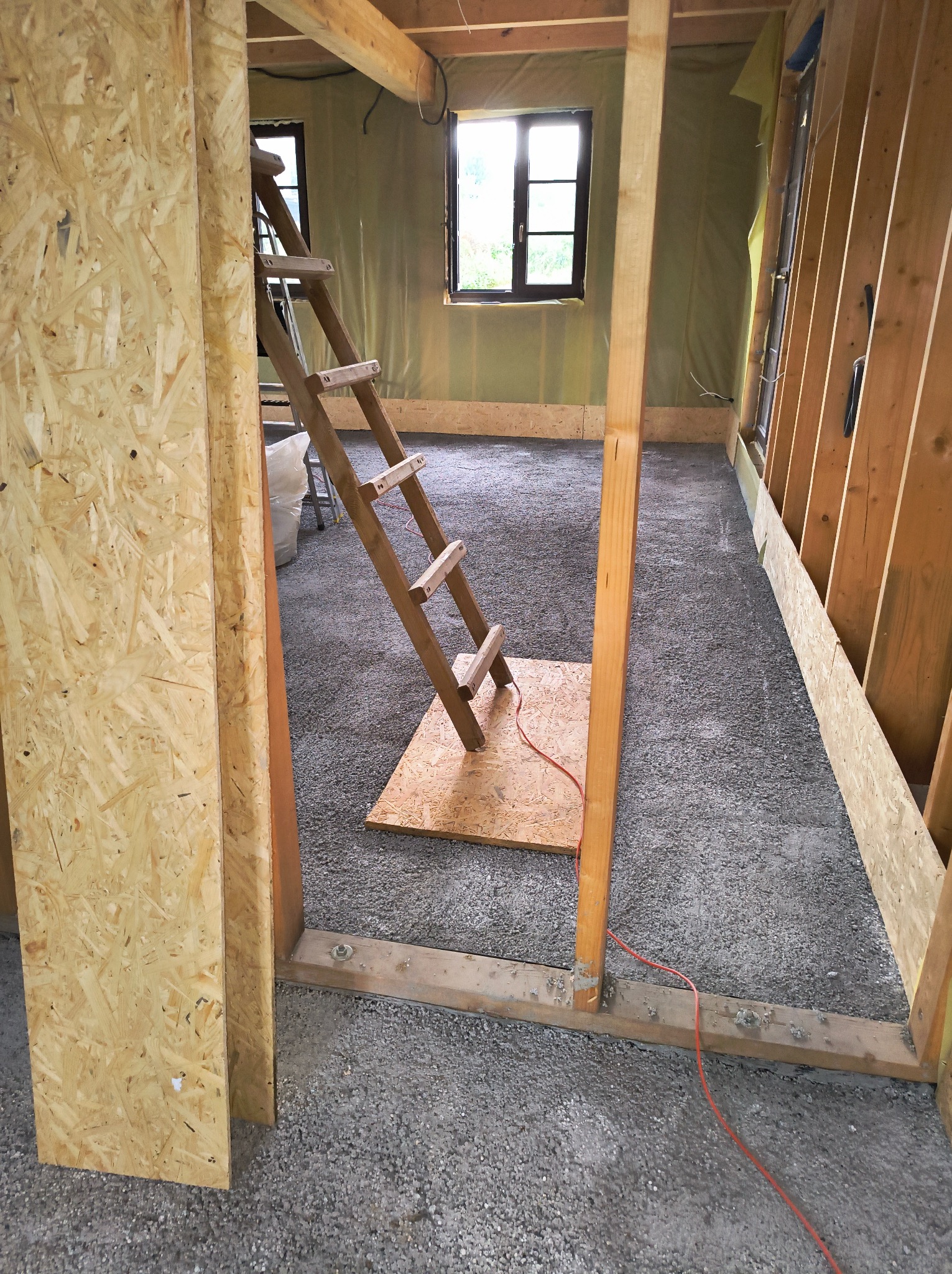 Wnętrze budynku w trakcie ocieplania ścian, widoczna drabina oparta o ścianę, szara podłoga, ściany w trakcie izolacji, drewniana konstrukcja szkieletowa i fragment płyty OSB.