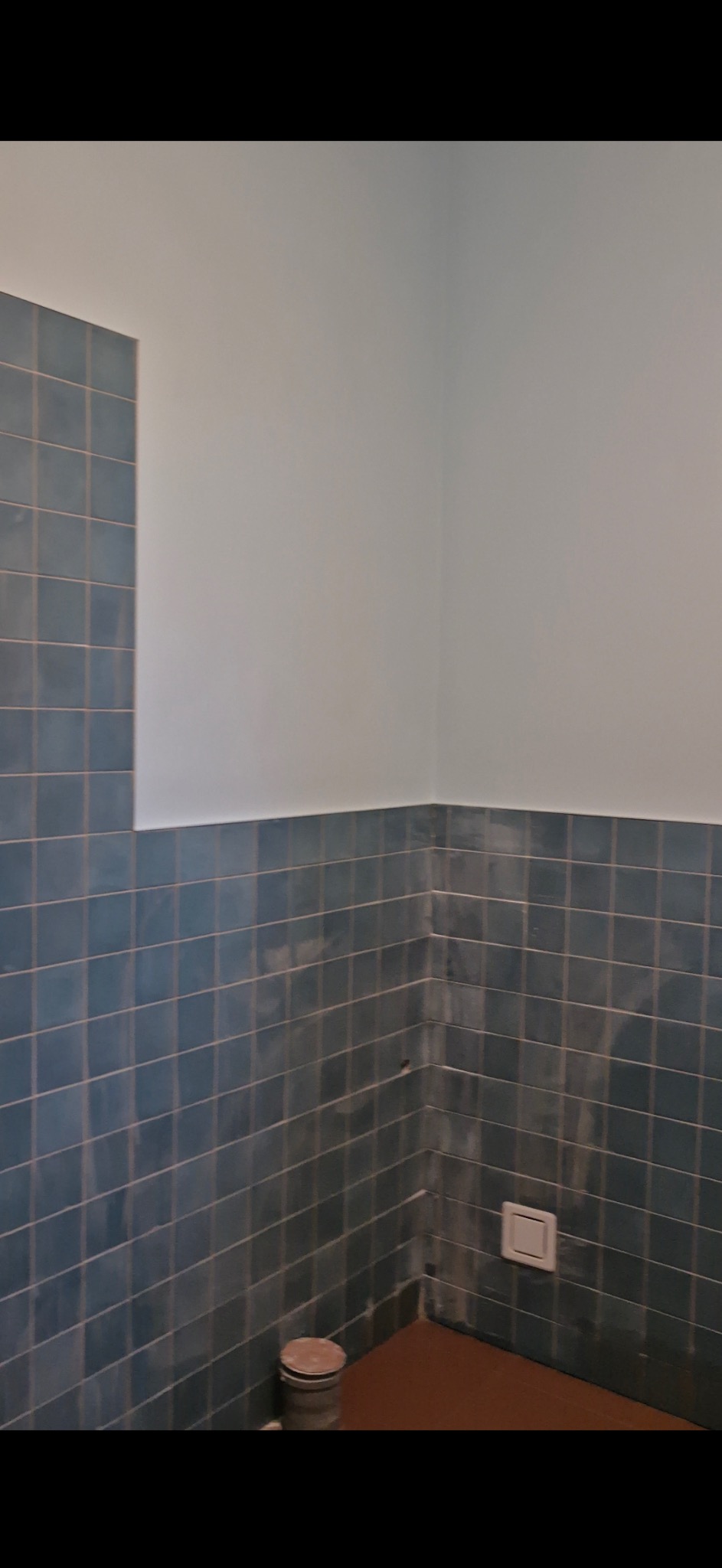 Narożnik łazienki z niebieskimi, kwadratowymi płytkami, częściowo pokryty białym tynkiem. Widoczne ślady po czyszczeniu i wiaderko z resztkami zaprawy na brązowej podłodze. W ścianie zamontowany...