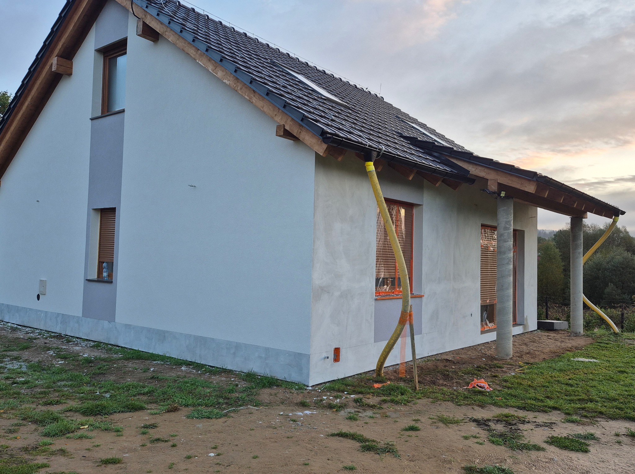 Surowy tynk elewacyjny na nowym domu jednorodzinnym, widoczne rury wentylacyjne, okna zabezpieczone folią, przygotowanie do dalszych prac wykończeniowych.