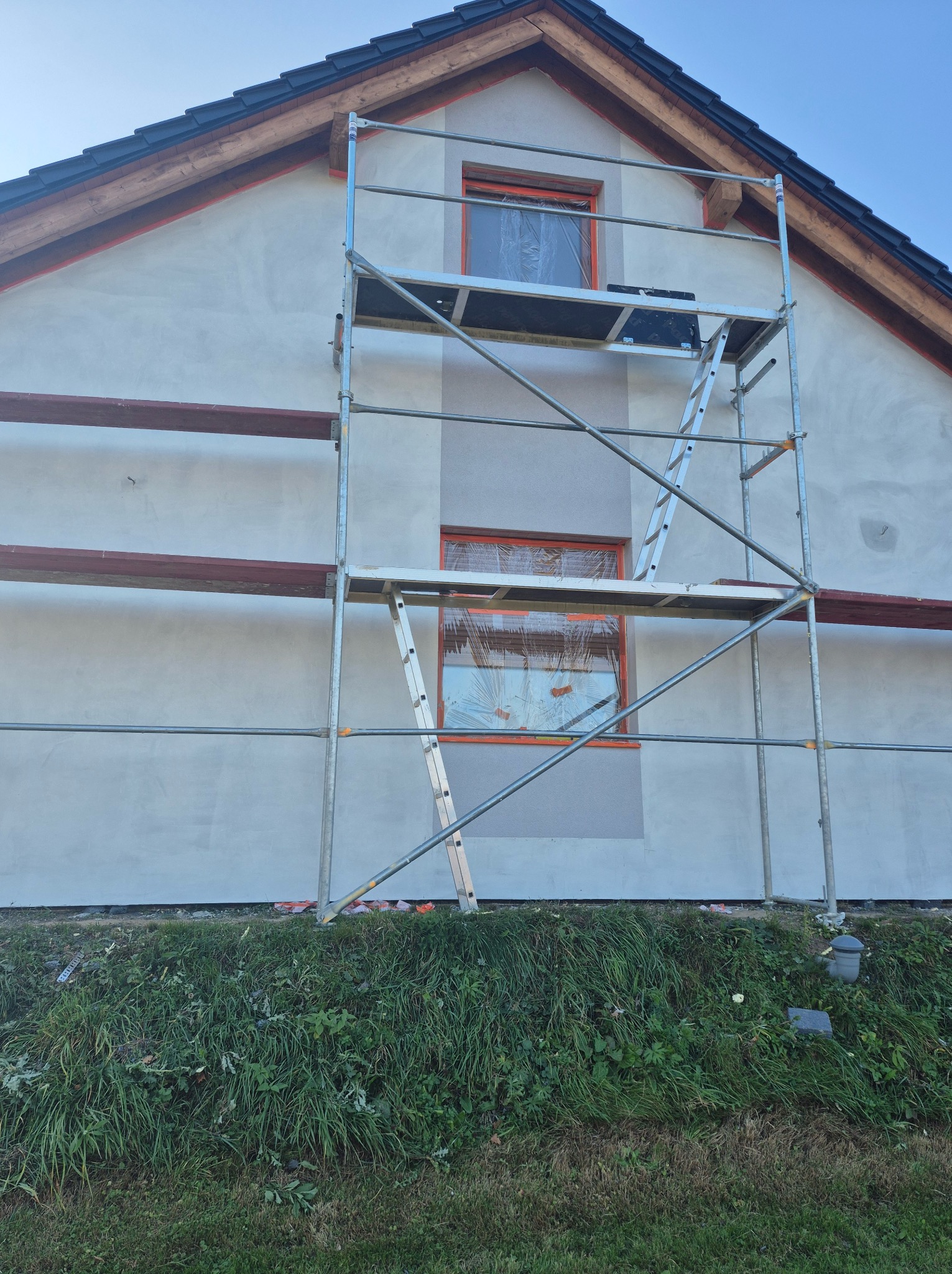 Dom jednorodzinny w trakcie prac elewacyjnych: świeżo nałożony tynk w jasnym odcieniu, okna zabezpieczone folią, rusztowanie metalowe z drabinką, widoczne pozostałości po deskach szalunkowych...