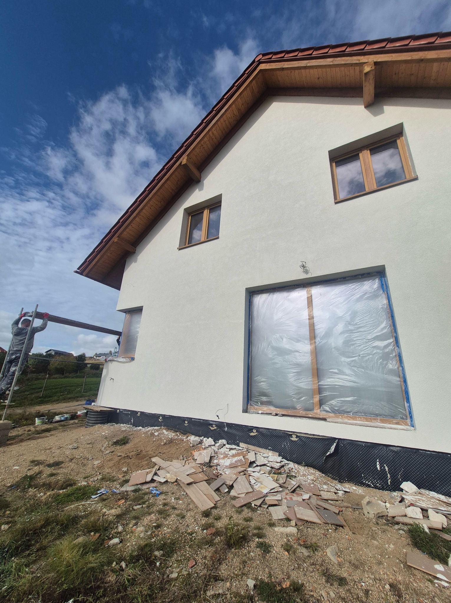 Świeżo otynkowana elewacja domu jednorodzinnego z drewnianą stolarką okienną, okna zabezpieczone folią, widoczne rusztowanie i pozostałości materiałów budowlanych na ziemi.