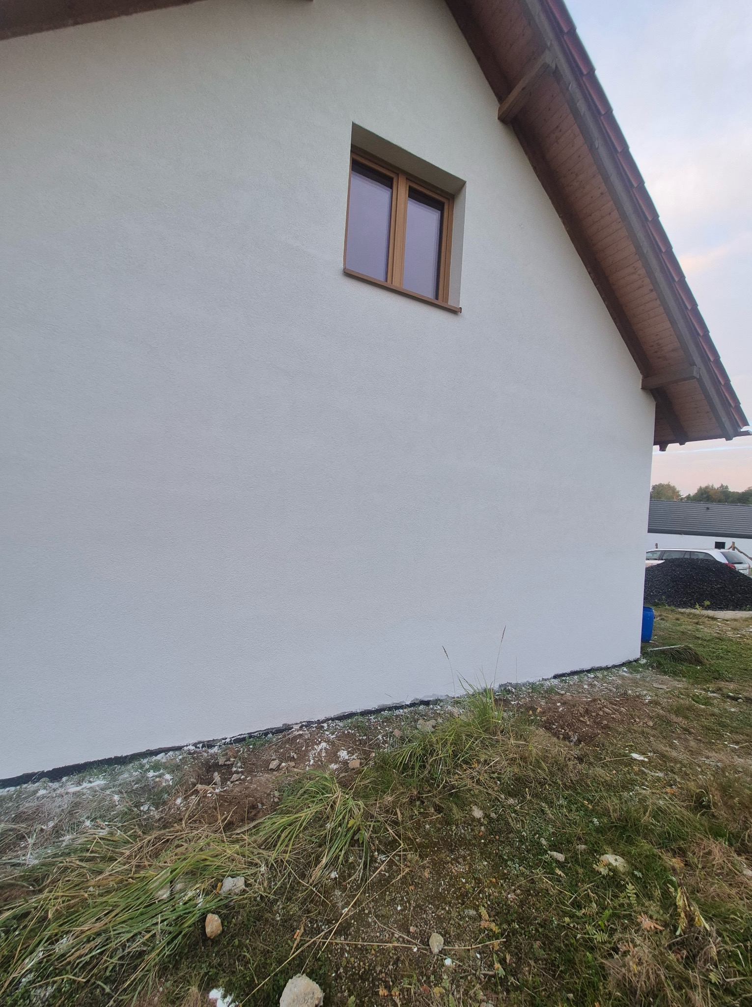 Boczny widok domu z białą elewacją, drewnianym oknem i dachem z drewnianą podbitką, trawa i gruz przy fundamencie.