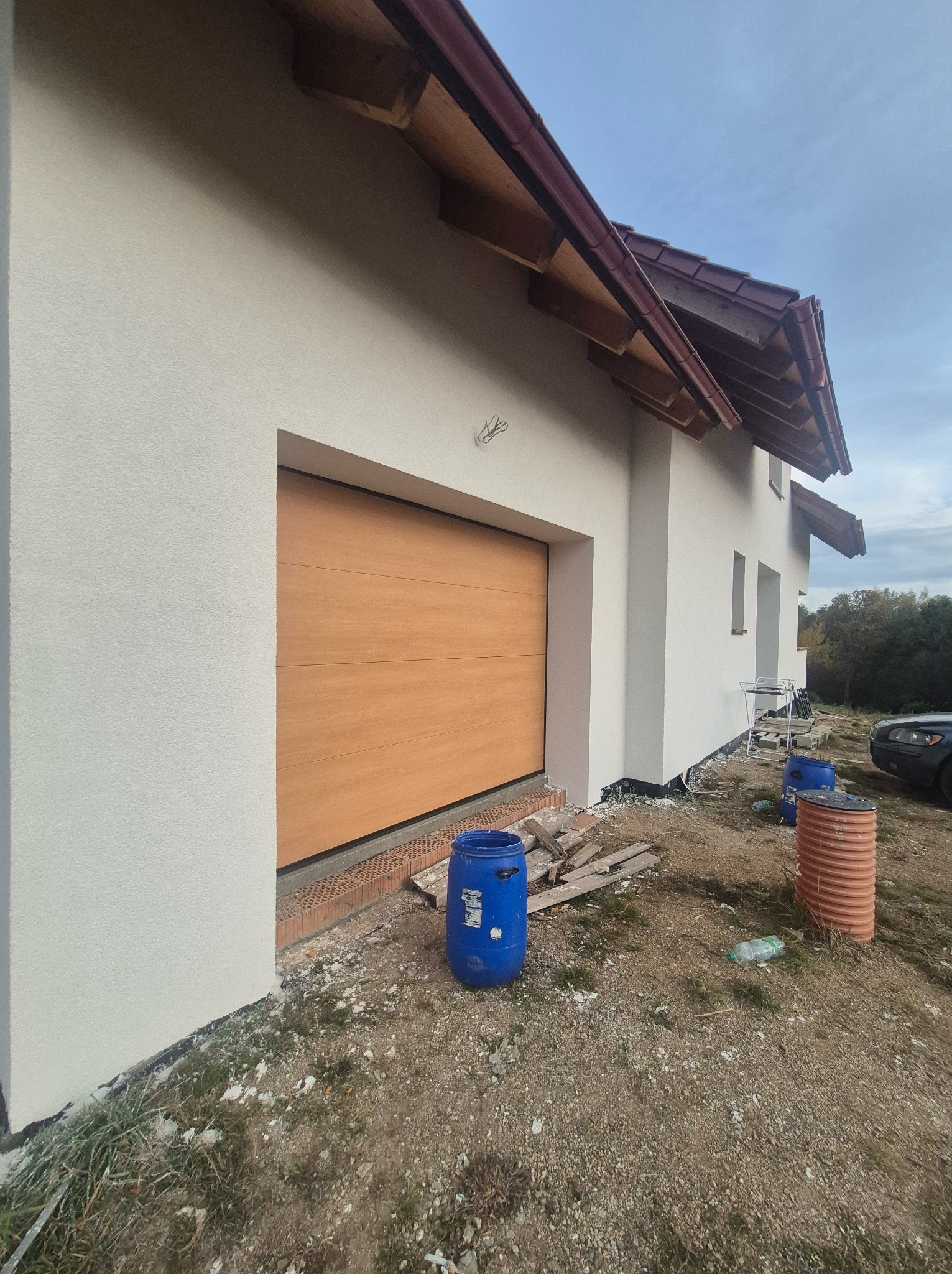Świeżo otynkowana biała elewacja domu z brązowym dachem, widoczna drewniana brama garażowa, niebieskie beczki i elementy budowlane na niezagospodarowanym terenie.