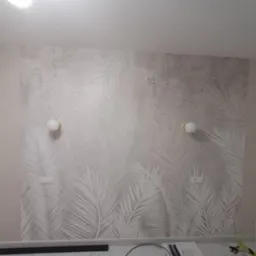 Ściana z tapetą w motywy liści palmowych, oświetlona dwiema okrągłymi lampami ściennymi z białymi kloszami, gniazdka elektryczne widoczne na tapecie.