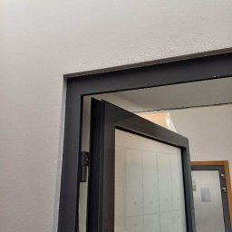 BudService Vasyl Kretsul - Częściowo otwarte, grafitowe, przeszklone drzwi wewnętrzne w jasnym pomieszczeniu. Widoczne zawiasy i fragment futryny. W tle kolejne drzwi.