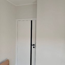 BudService Vasyl Kretsul - Minimalistyczne wnętrze: białe ściany, listwa przypodłogowa i nowoczesne drzwi z czarnym akcentem. Widoczny fragment kartonu w rogu pomieszczenia.