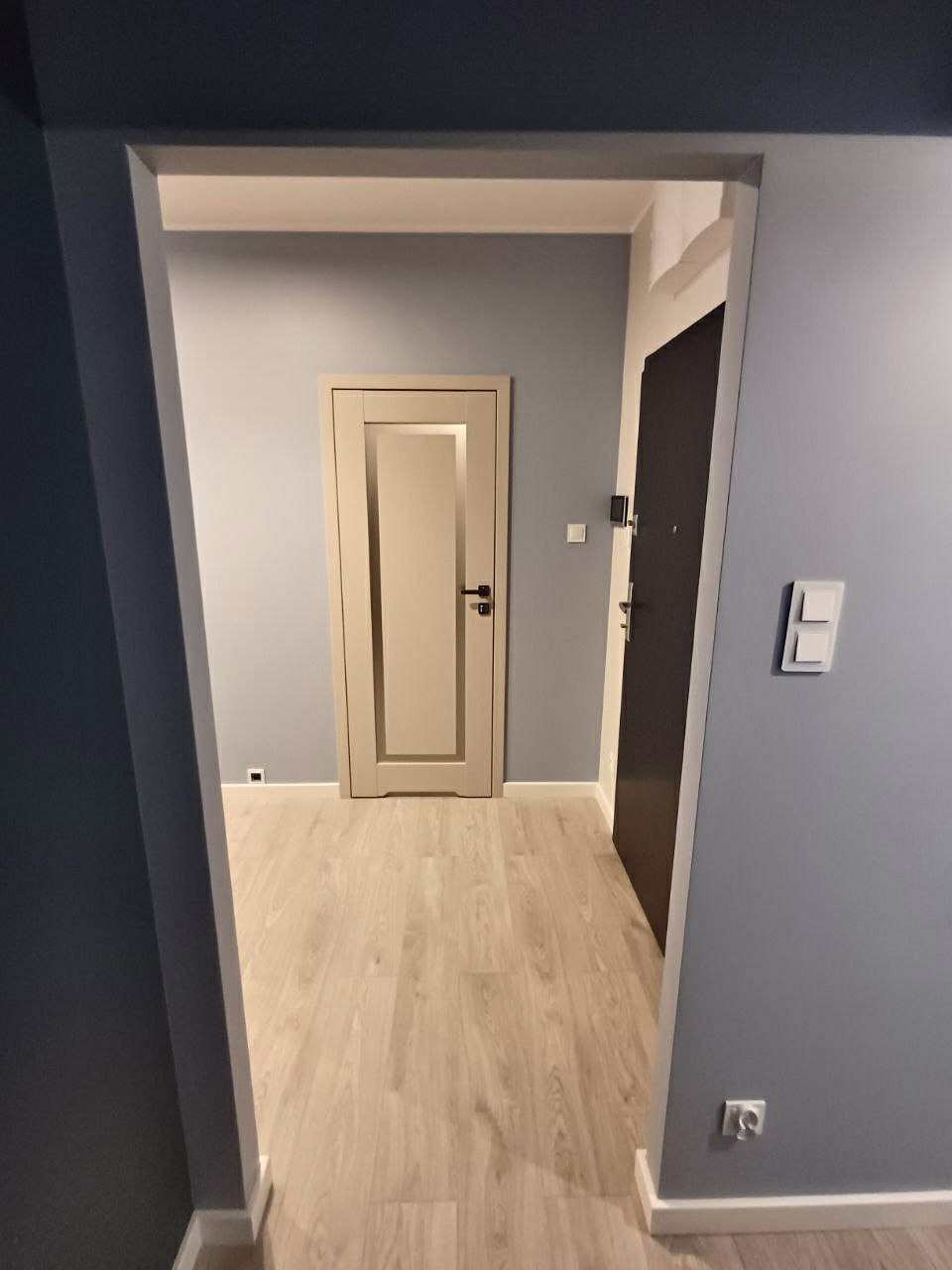 Jasna podłoga w przedpokoju z szarymi ścianami i dwiema parami drzwi w odcieniach beżu i ciemnego brązu. Widoczne gniazdka i włączniki światła.