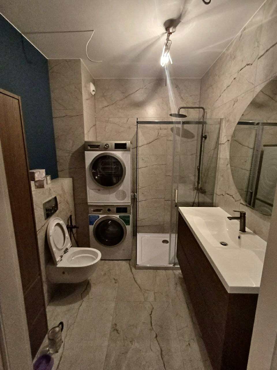 Wykończona łazienka z marmurowymi płytkami, kabiną prysznicową, umywalką z szafką w kolorze ciemnego drewna i pralko-suszarką w kolumnie. Toaleta podwieszana z otwartą klapą.