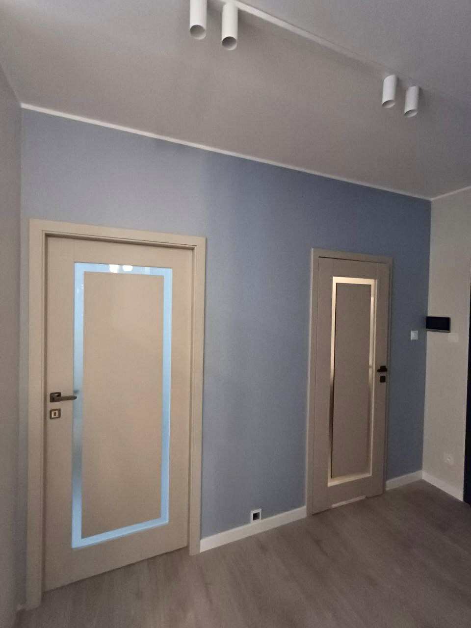 Dwa odcienie szarości na ścianie z dwiema parami drzwi wewnętrznych w odcieniach beżu i szarości. Podłoga laminowana. Oświetlenie sufitowe punktowe.