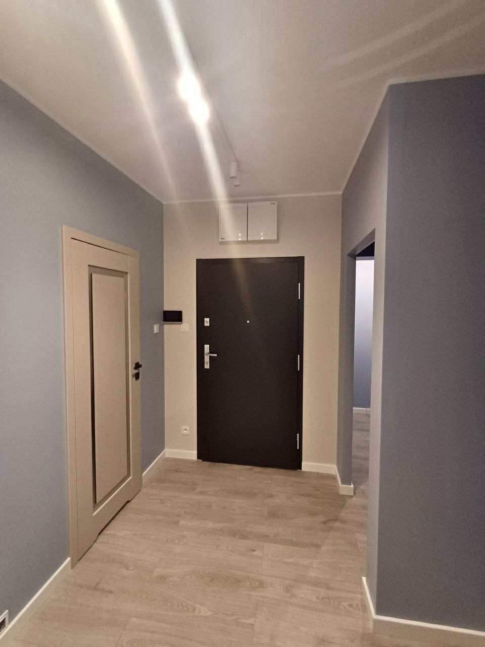 Wykończony korytarz w odcieniach szarości z drewnopodobną podłogą i dwiema parami drzwi. Jedne drzwi są otwarte.