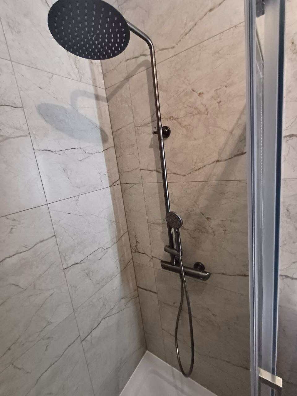 Nowoczesny prysznic z deszczownicą i baterią w kolorze grafitowym na tle płytek imitujących marmur w wykończonej łazience.