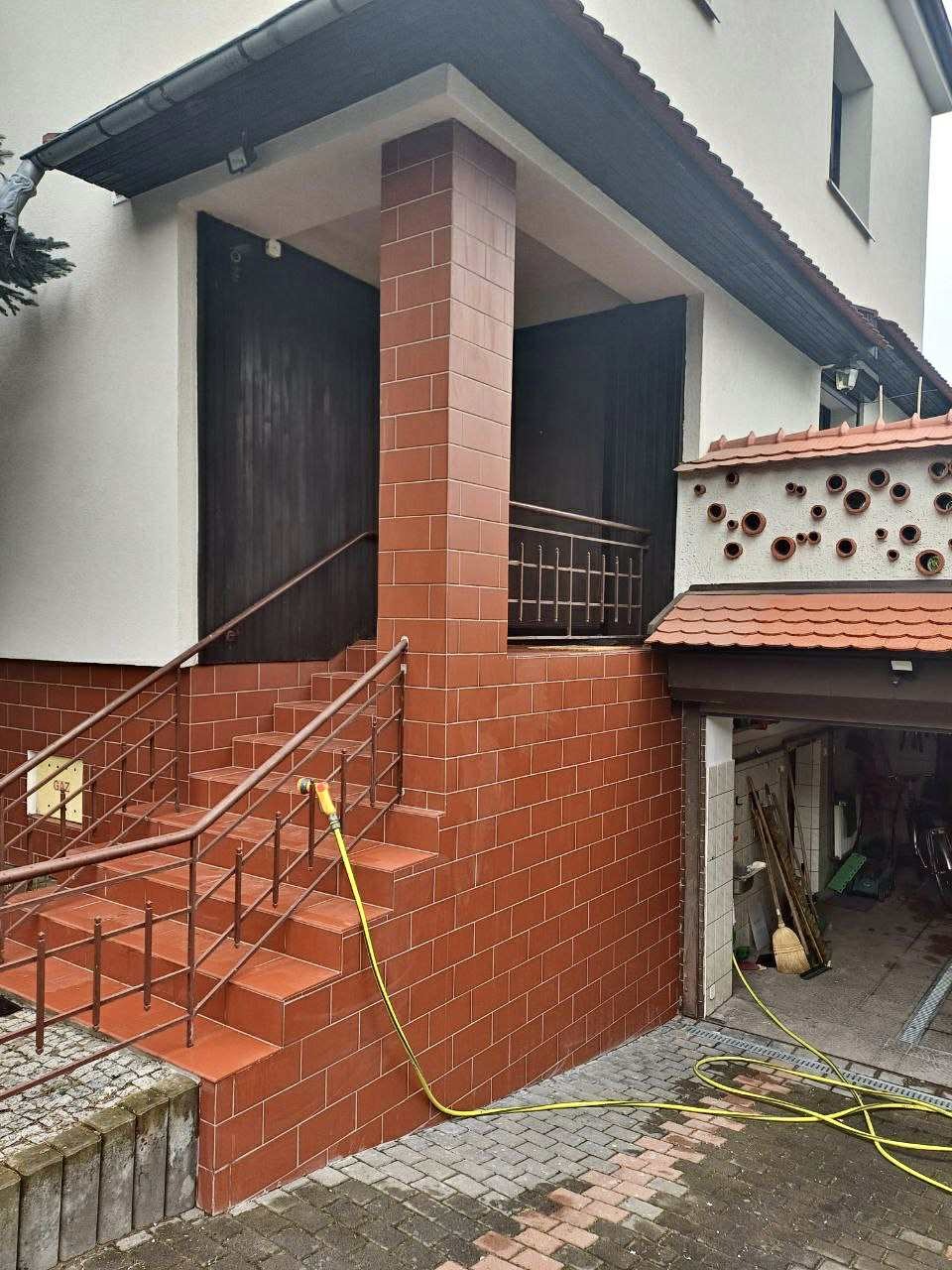 Klatka schodowa i kolumna wejściowa wyłożone czerwoną, błyszczącą terakotą. Żółty wąż ogrodowy leży na brukowanej kostce. Widoczny garaż z narzędziami.