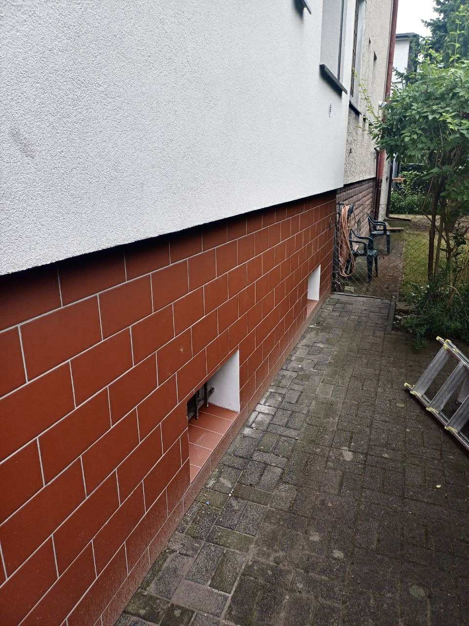 Wykończenie elewacji domu z czerwoną cegłą klinkierową na cokole, widoczne okienko piwniczne i fragment chodnika z kostki brukowej. Ujęcie z boku budynku.