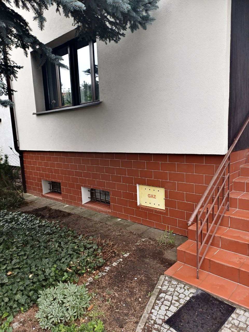 Elewacja domu z oknem, czerwoną cegłą klinkierową w dolnej części i schodami wejściowymi. Tabliczka gazowa na ścianie.