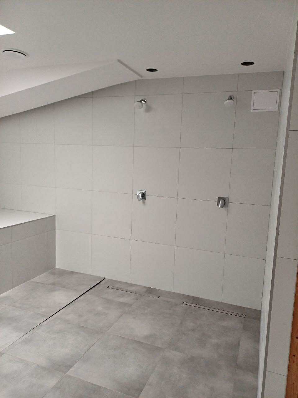 Wykończona łazienka na poddaszu z szarymi płytkami na ścianach i podłodze, minimalistyczna armatura, odpływ liniowy. Widoczne skosy sufitu.