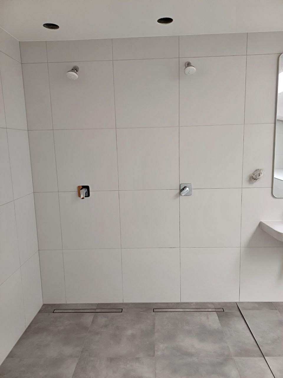 Nowoczesna łazienka z białymi, kwadratowymi płytkami na ścianach, szarymi płytkami podłogowymi i minimalistycznymi odpływami liniowymi. Dwa prysznice sufitowe.