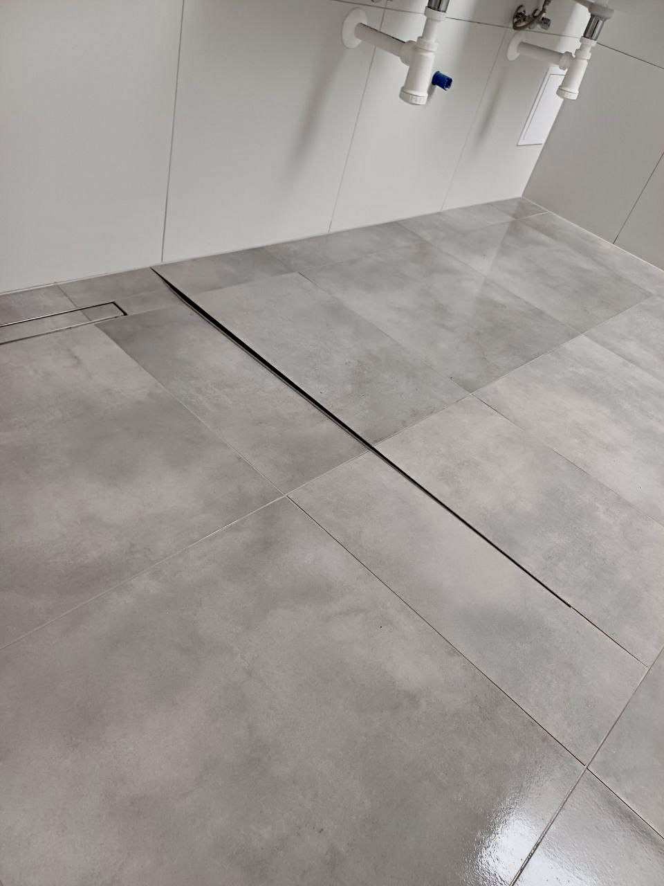 Szare, matowe płytki podłogowe w łazience, z widocznym liniowym odpływem. Minimalistyczny design, jasne ściany i fragment instalacji sanitarnej w tle.