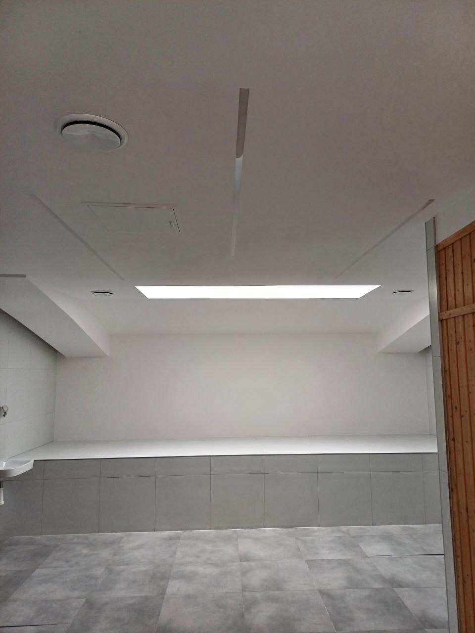 Wykończona łazienka z szarymi płytkami na podłodze i ścianach. Sufit podwieszany z oświetleniem LED. Widok na minimalistyczne wnętrze z elementami drewna.