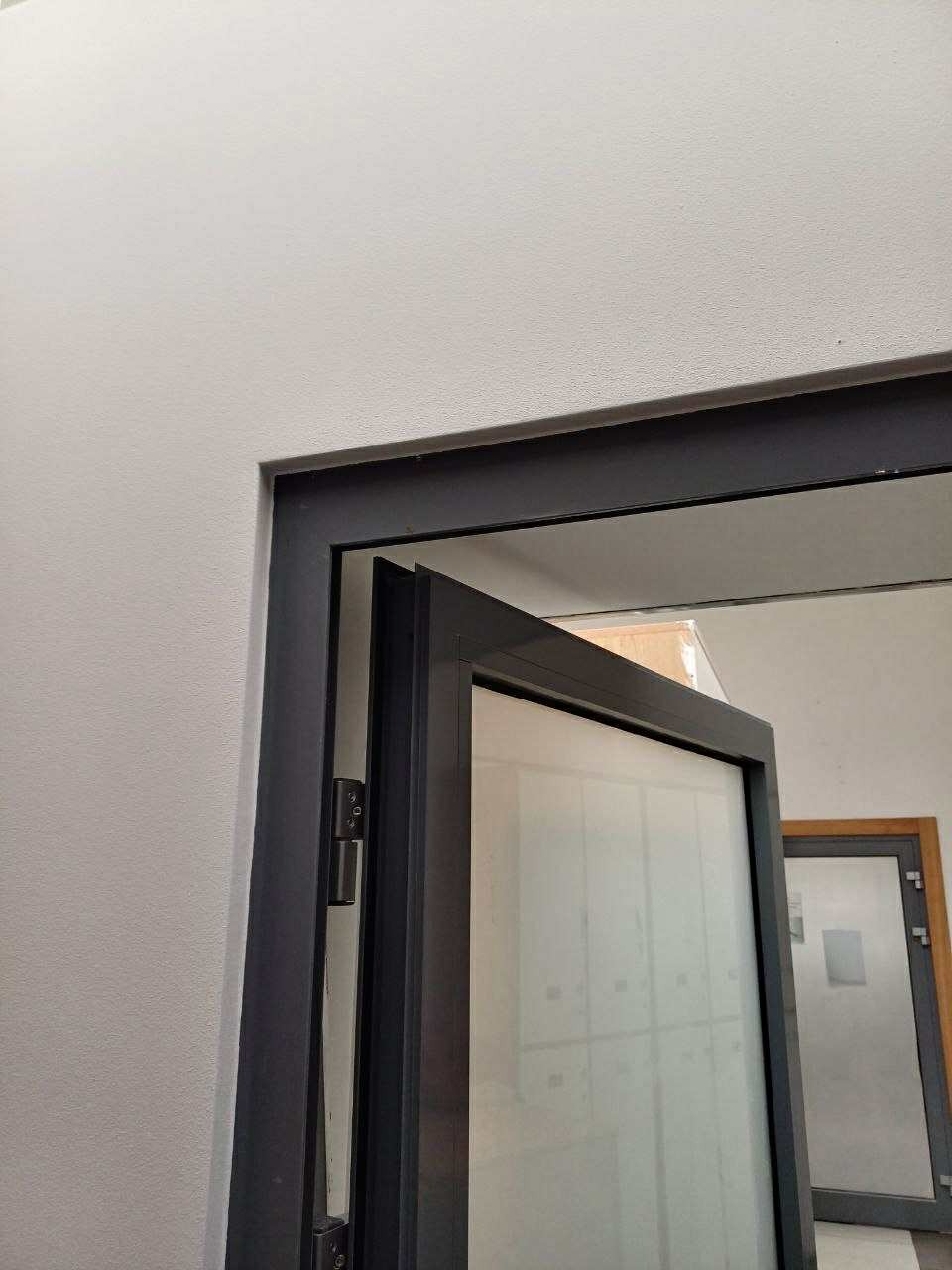 Częściowo otwarte, grafitowe, przeszklone drzwi wewnętrzne w jasnym pomieszczeniu. Widoczne zawiasy i fragment futryny. W tle kolejne drzwi.