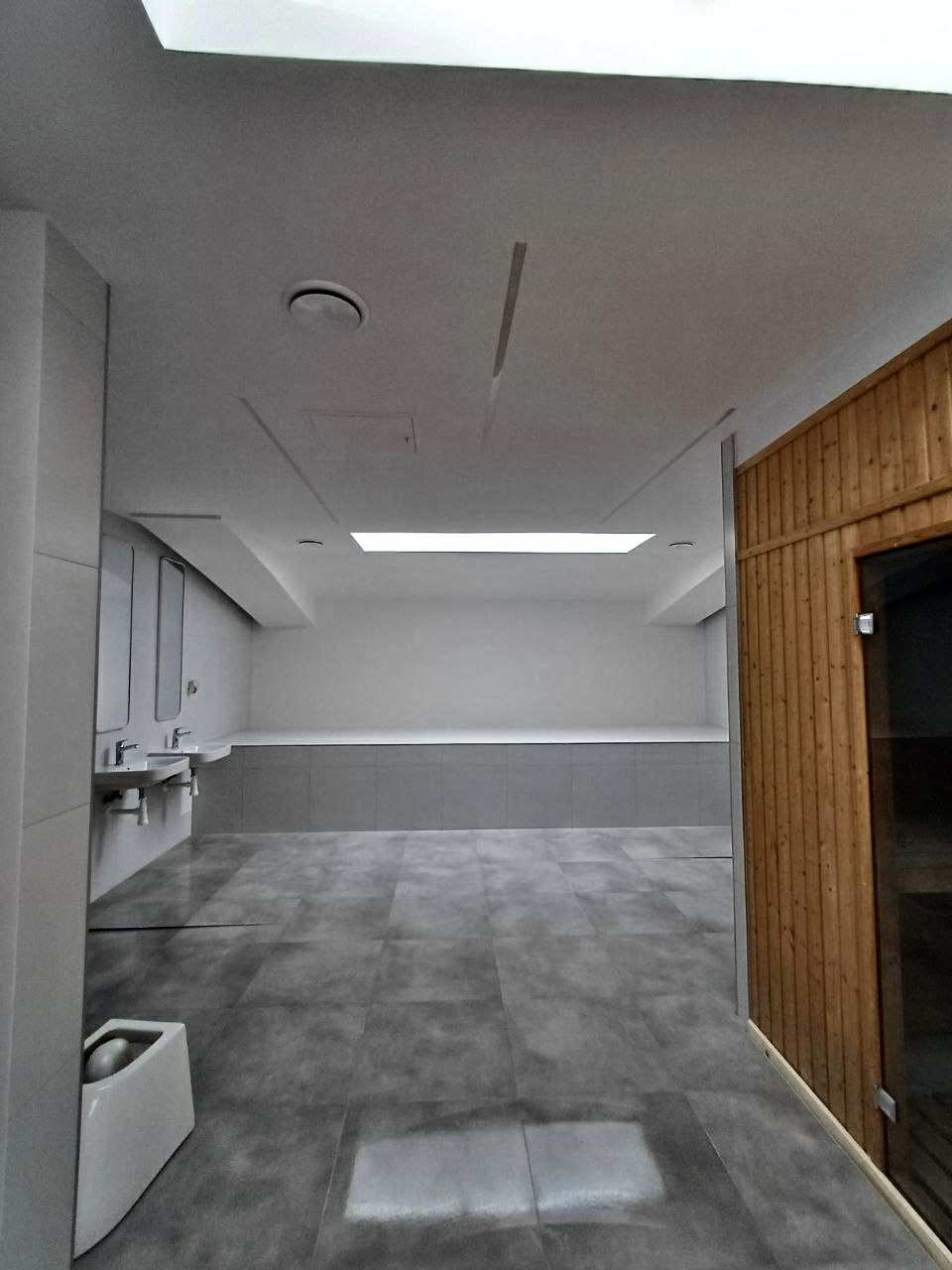 Nowoczesna łazienka publiczna z szarą podłogą, umywalkami i drewnianą sauną. Minimalistyczny design, oświetlenie sufitowe.