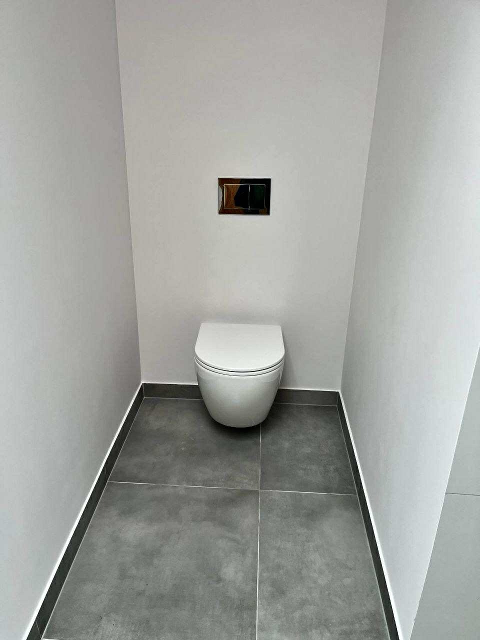 Minimalistyczna toaleta w szarościach: podwieszana miska WC, ciemne płytki podłogowe, chromowany przycisk spłukujący. Nowoczesne wykończenie małej łazienki.