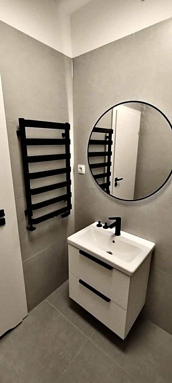 Nowoczesna łazienka w szarościach z czarnymi akcentami: okrągłe lustro, grzejnik drabinkowy i biała szafka z umywalką. Minimalistyczny design, płytki ceramiczne na ścianach.