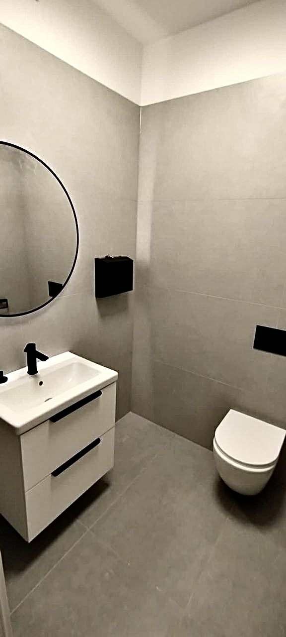 Minimalistyczna łazienka w szarościach: okrągłe lustro, biała szafka z umywalką, podwieszana toaleta i czarne akcenty. Nowoczesne wykończenie.