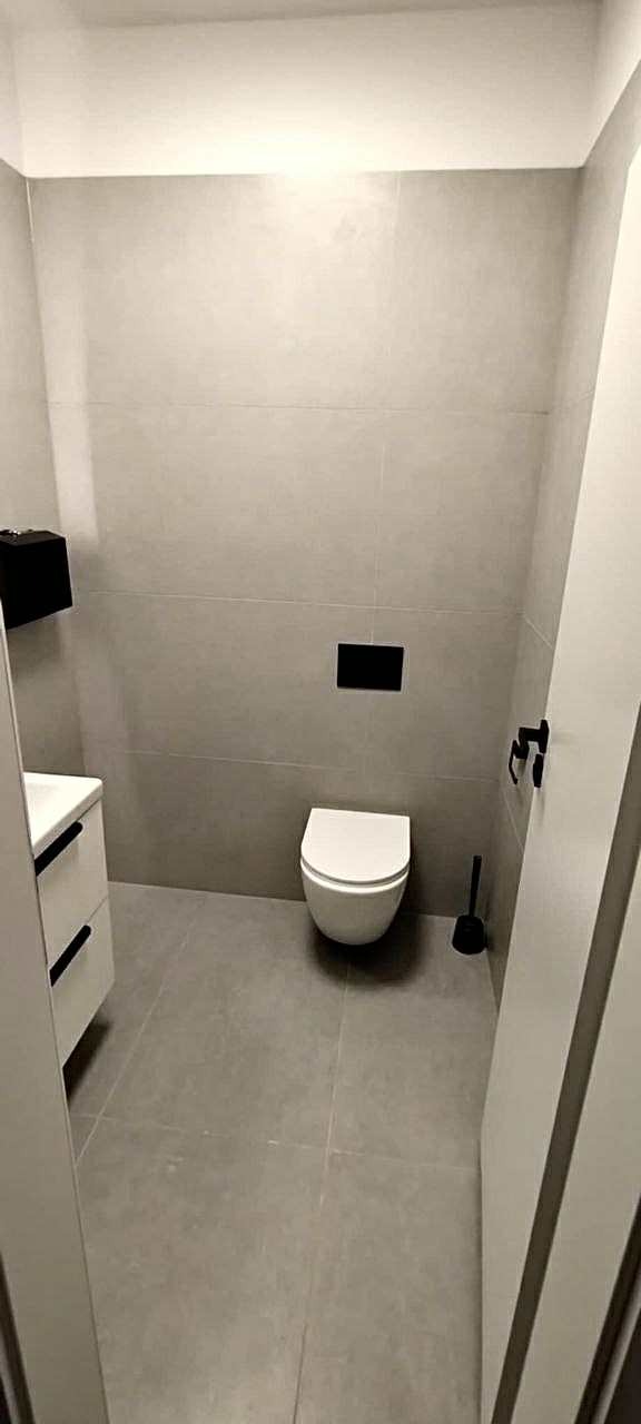 Minimalistyczna łazienka po remoncie: szare płytki, podwieszana toaleta z czarnym przyciskiem, biała szafka z czarnymi uchwytami i czarne akcesoria.