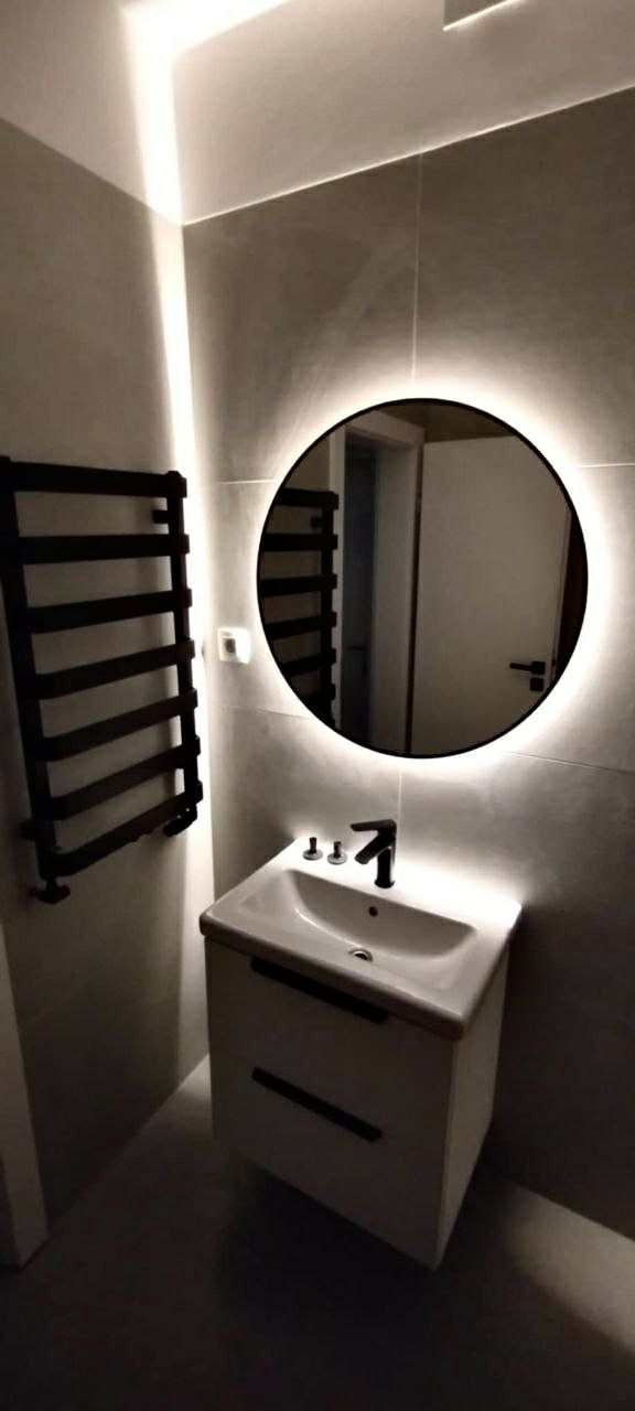 Nowoczesna łazienka z okrągłym lustrem LED, umywalką na szafce z ciemnymi uchwytami i czarnym grzejnikiem drabinkowym na szarej ścianie.