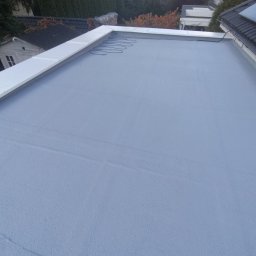 M&M Mariusz Sitek - Szary, płaski dach pokryty jednolitą powłoką, z widocznymi przewodami grzewczymi i białym obramowaniem, na tle zieleni drzew i zabudowań.