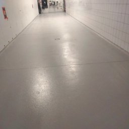 M&M Mariusz Sitek - Gładka, szara posadzka żywiczna w długim, korytarzu przemysłowym z białymi, kafelkowymi ścianami, widoczne łączenia segmentów podłogi i kratka ściekowa.