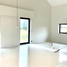 M&M Mariusz Sitek - Wnętrze domu z wysokim sufitem i dużymi oknami, świeżo pomalowane na biało, z widocznym wałkiem do malowania podłogi i kuwetą z farbą w rogu