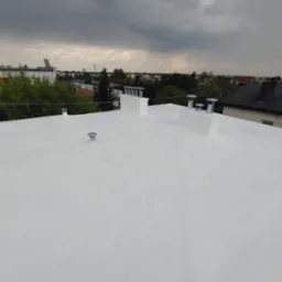 Biały, płaski dach po renowacji w Kaliszu, widok z góry. Kominy i wentylatory na tle pochmurnego nieba i zabudowań miejskich. Nowa izolacja dachu.