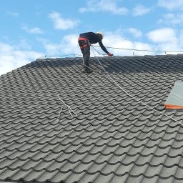 M&M Mariusz Sitek - Pracownik w kasku i uprzęży zabezpieczającej na szarym dachu z dachówki ceramicznej, podczas montażu zabezpieczeń. Błękitne niebo w tle.