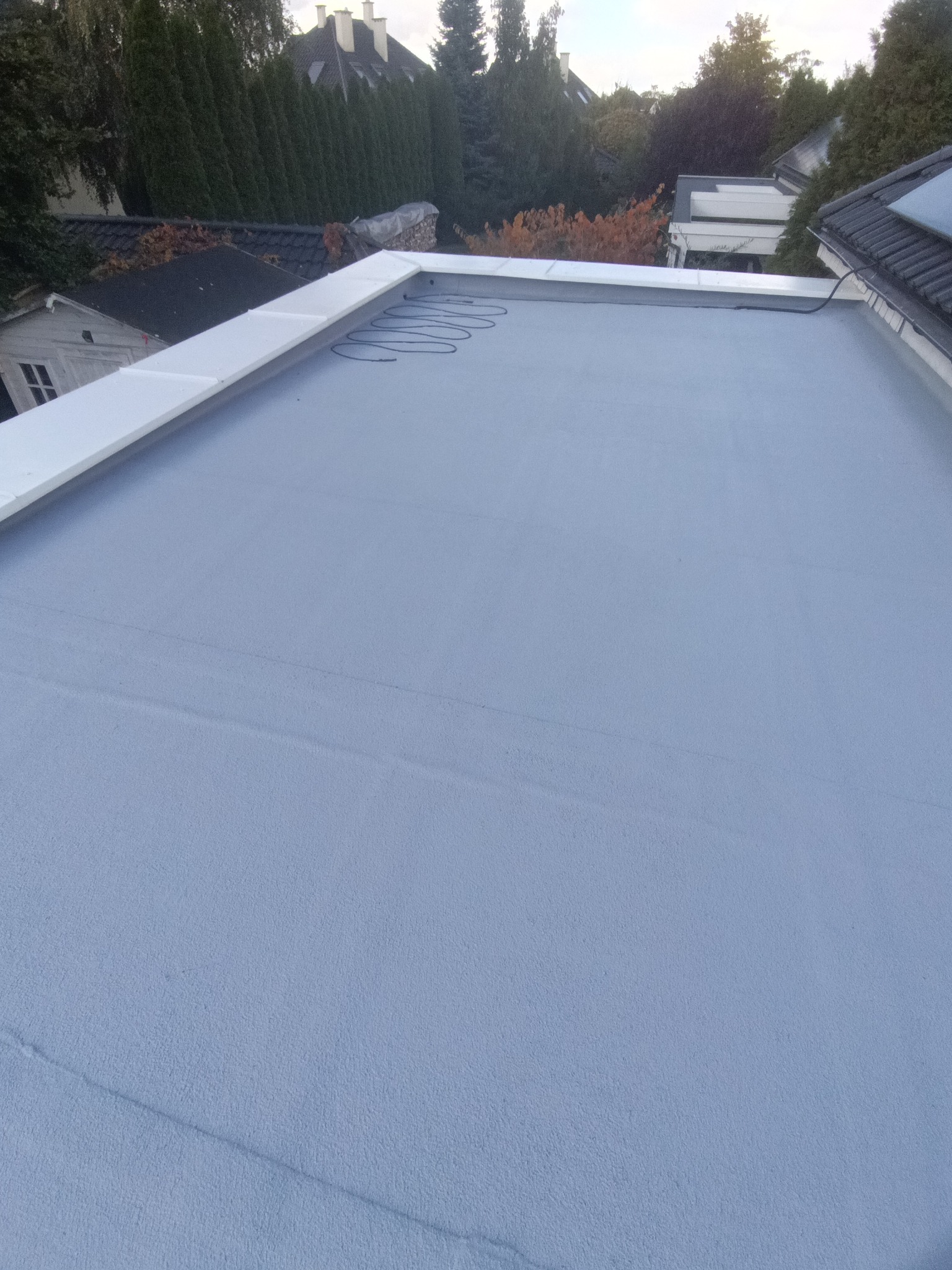 Szary, płaski dach pokryty jednolitą powłoką, z widocznymi przewodami grzewczymi i białym obramowaniem, na tle zieleni drzew i zabudowań.