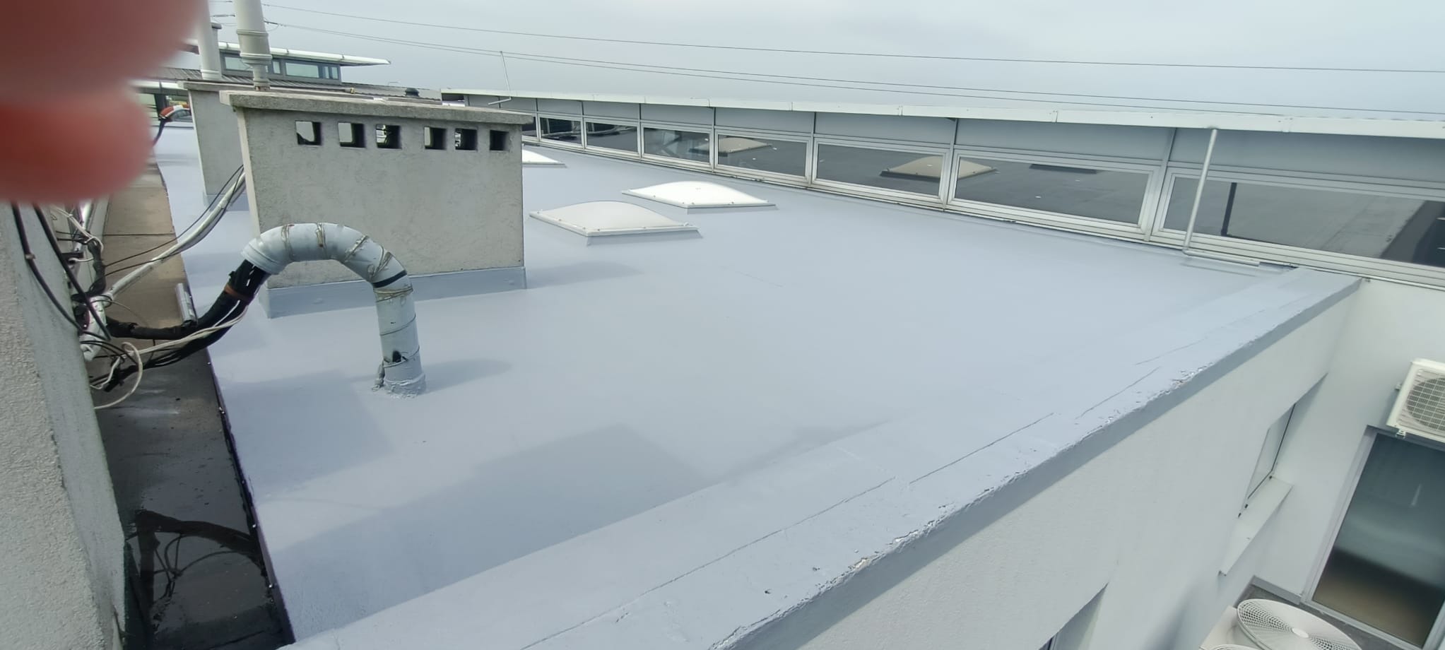 Szary dach pokryty jednolitą, gładką powłoką, z elementami wentylacyjnymi i świetlikami, widoczne okna budynku w tle.