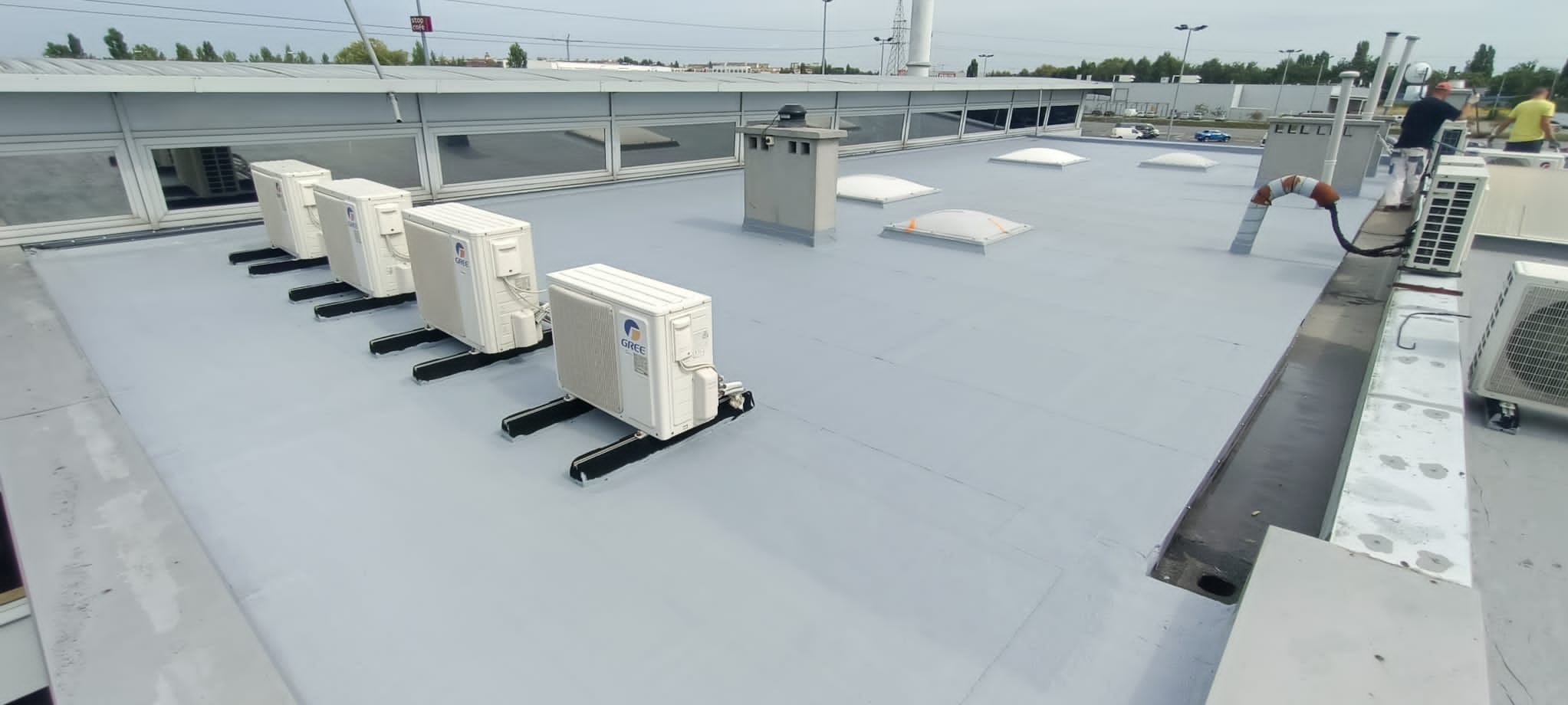 Szary dach pokryty jednolitą powłoką, z rzędem klimatyzatorów i pracownikami w tle, prawdopodobnie po nałożeniu warstwy ochronnej.