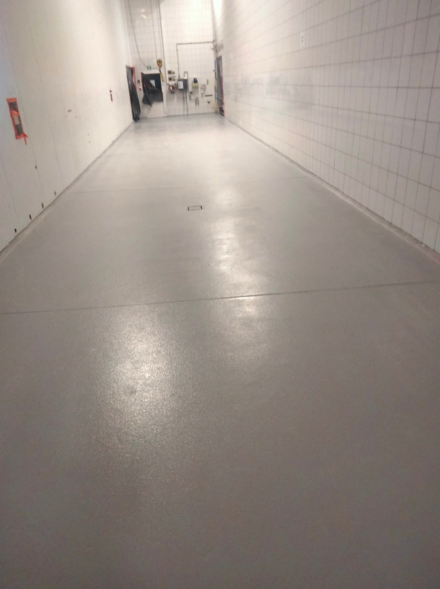 Gładka, szara posadzka żywiczna w długim, korytarzu przemysłowym z białymi, kafelkowymi ścianami, widoczne łączenia segmentów podłogi i kratka ściekowa.