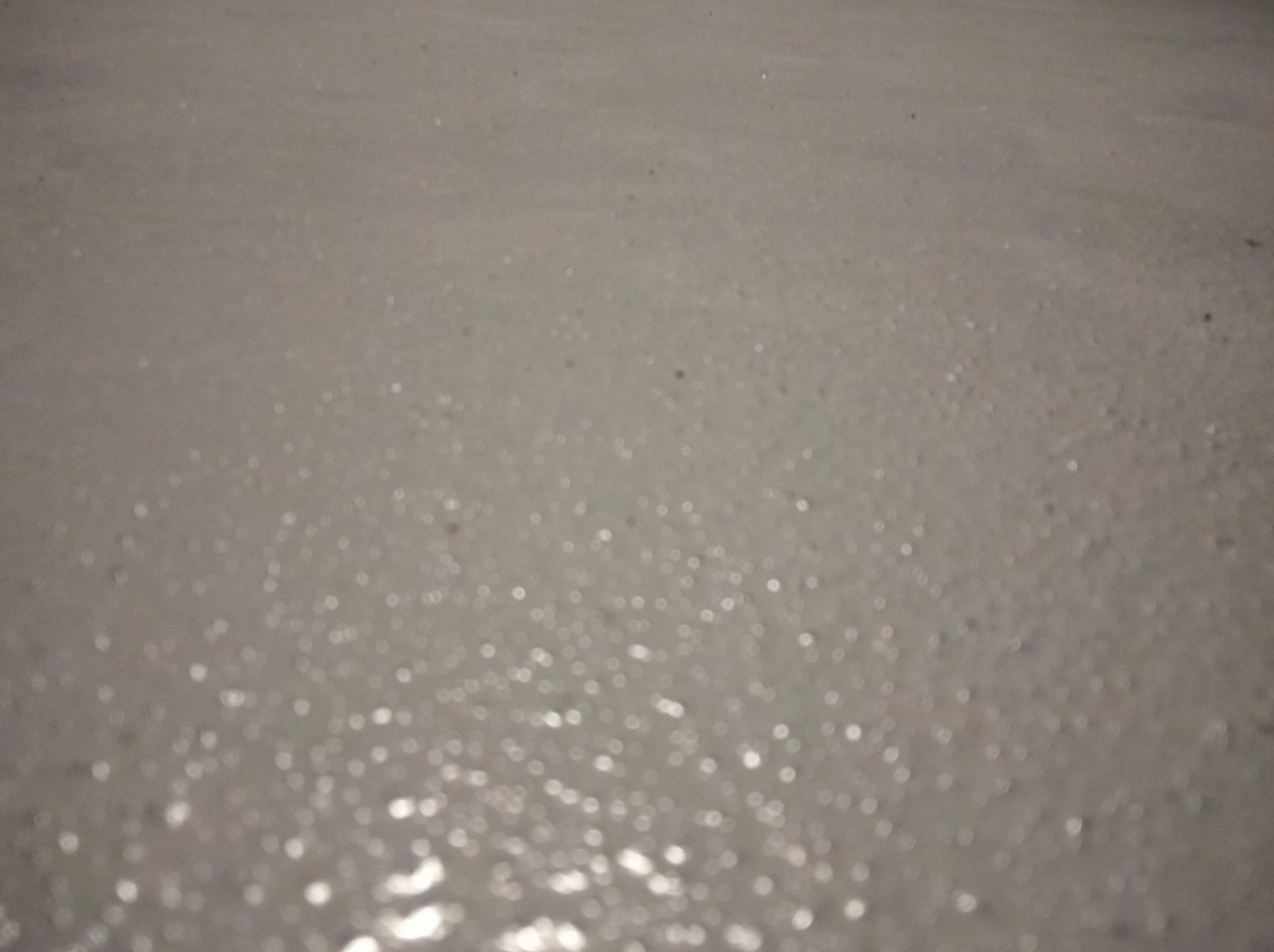 Niejasne zbliżenie na szarą, błyszczącą powierzchnię, prawdopodobnie świeżo wylaną i mokrą, z drobnymi refleksami światła.