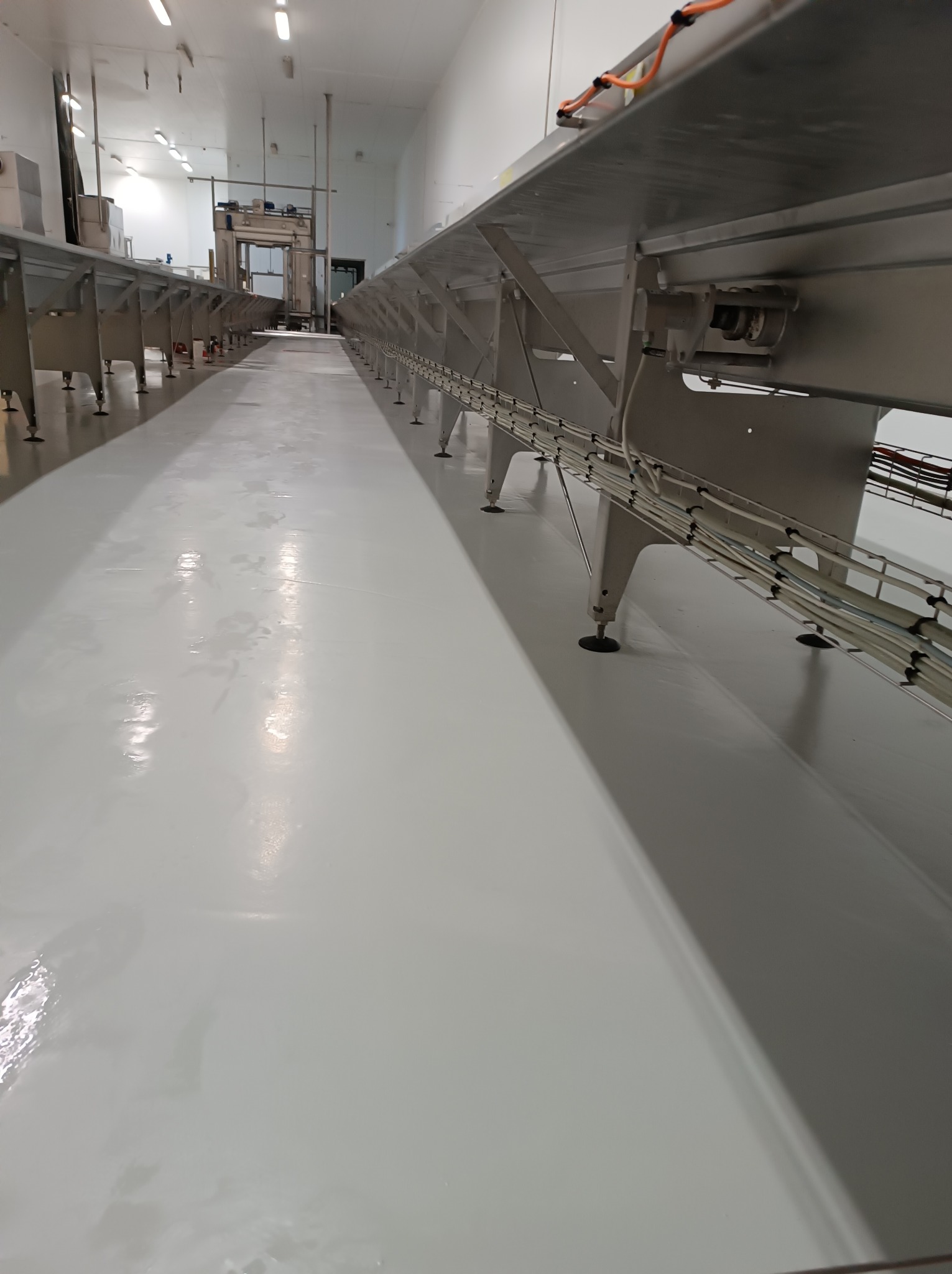 Biała, błyszcząca posadzka w hali produkcyjnej z metalowymi konstrukcjami podtrzymującymi system transportowy, widok z perspektywy podłogi.