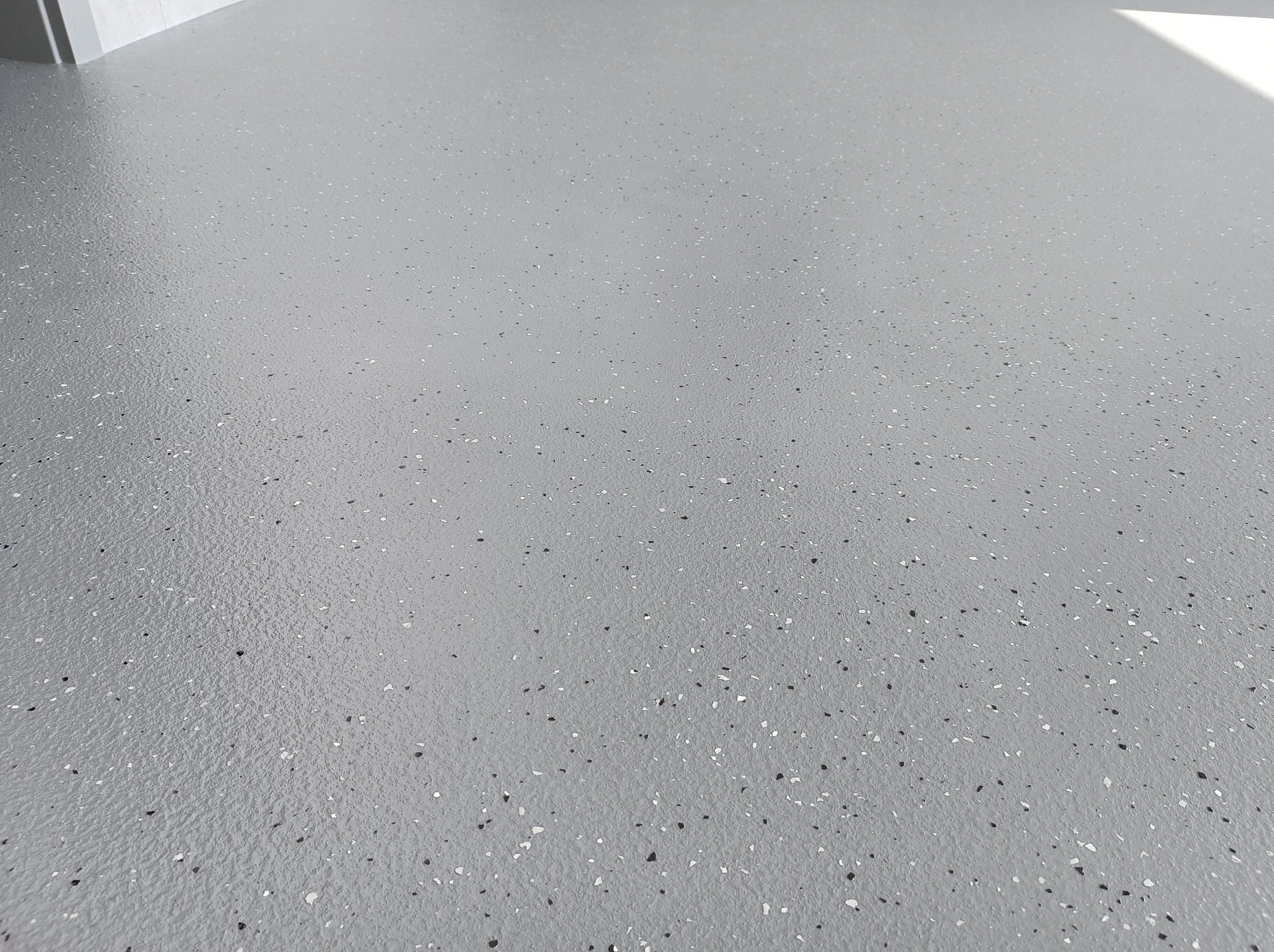 Szara, gładka posadzka z drobnymi, białymi i czarnymi wtrąceniami, widoczna z perspektywy pod lekkim kątem, oświetlona naturalnym światłem.