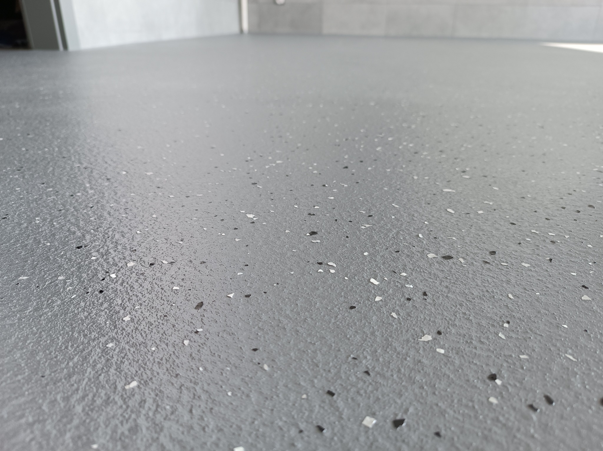 Szara, gładka posadzka z drobnymi, nieregularnymi, białymi i czarnymi wtrąceniami, widziana z niskiej perspektywy, ukazująca jej jednolitą strukturę i połysk.