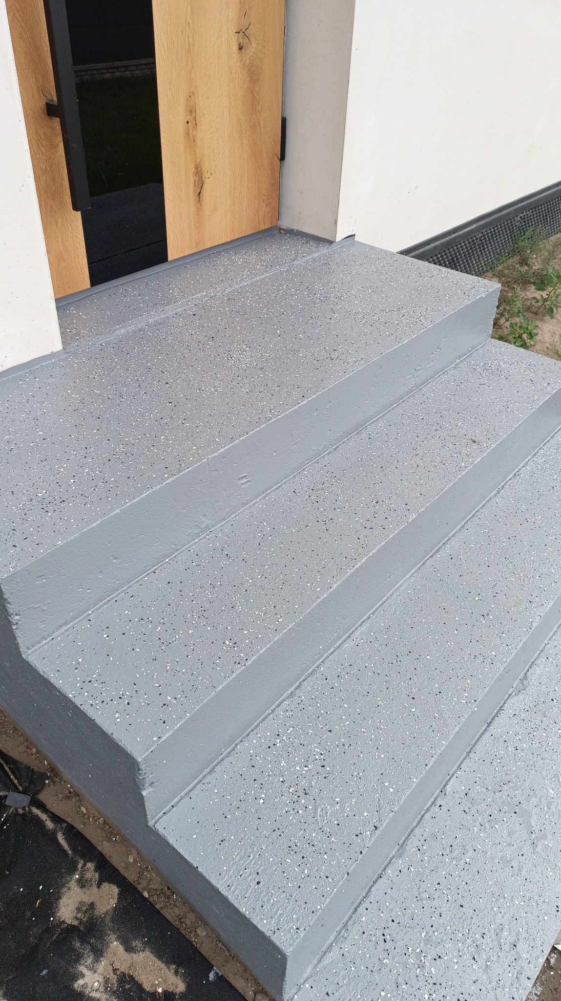 Zewnętrzne schody wejściowe pokryte szarą posadzką z drobnymi, ciemnymi i jasnymi plamkami, prowadzące do drewnianych drzwi wejściowych z czarną klamką.