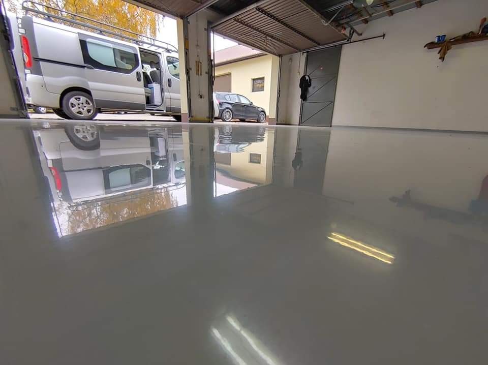 Błyszcząca, szara posadzka żywiczna w garażu, odbijająca zaparkowany samochód i otwarte drzwi wjazdowe.